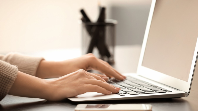 Attēlā redzamas rokas, kas raksta uz portatīvā datora tastatūras. Uz galda blakus datoram atrodas arī viedtālrunis un pildspalvu turētājs ar pildspalvām.